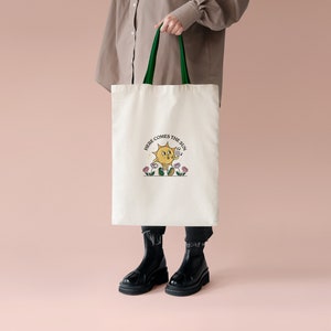 May include: Cream tote bag with green handles. The bag has a cartoon sun illustration with the text "HERE COMES THE SUN." The sun is smiling and holding a musical note. The bag is held against a pink background.