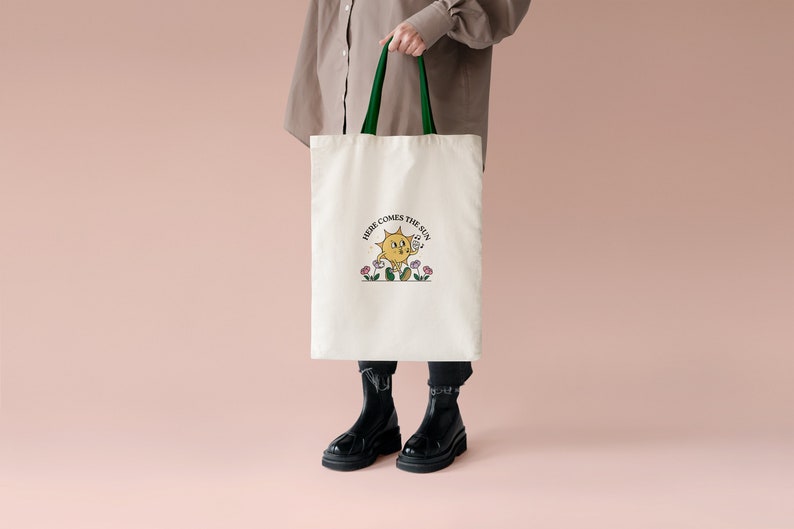 May include: Cream tote bag with green handles. The bag has a cartoon sun illustration with the text "HERE COMES THE SUN." The sun is smiling and holding a musical note. The bag is held against a pink background.