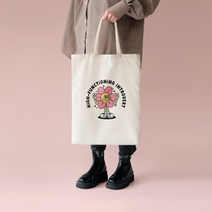 May include: A white tote bag with a pink flower graphic and the text "High-Functioning Introvert".