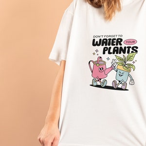 May include: White t-shirt with a graphic of a watering can and a potted plant with the text "Don't forget to water your plants".