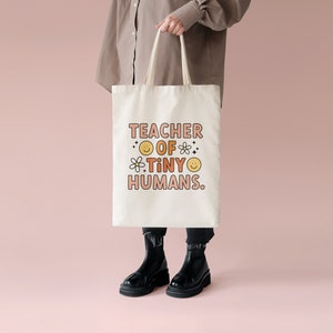 May include: A white tote bag with the text "TEACHER OF TINY HUMANS." in pink and orange lettering. The text is surrounded by small yellow smiley faces and flowers.