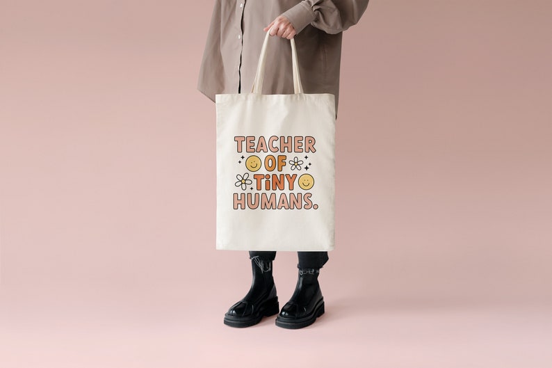May include: A white tote bag with the text "TEACHER OF TINY HUMANS." in pink and orange lettering. The text is surrounded by small yellow smiley faces and flowers.