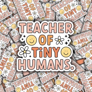 May include: A sticker with the text "Teacher of Tiny Humans." The text is in pink, orange, and yellow with a white outline. There are small white flowers and yellow smiley faces around the text.