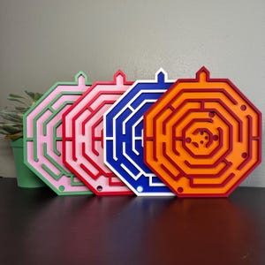 Op de afbeelding: Vier achthoekige doolhofpuzzels op een rij. Elke puzzel heeft een eigen kleurenschema: lichtgroen en roze, rood en lichtroze, blauw en wit, en oranje en donkerrood. Elk doolhof heeft een klein lipje aan de bovenkant en ronde gaten.