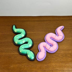 Puede incluir: Dos rompecabezas con forma de serpiente, uno verde y otro rosa, sobre una superficie de madera. Los rompecabezas están hechos de piezas que se encajan.