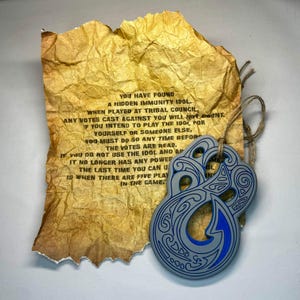May include: A gray and blue pendant with a tribal design hangs next to a piece of aged paper. The paper has text that reads, "YOU HAVE FOUND A HIDDEN IMMUNITY IDOL." The pendant is attached with twine. The paper is crumpled and torn.