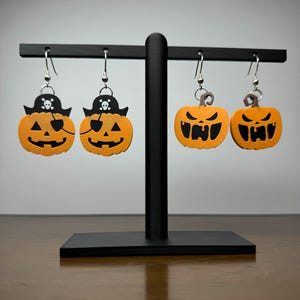 May include: Four Halloween-themed pumpkin earrings. Two earrings feature a pumpkin with a pirate hat and eye patch. The other two earrings feature a pumpkin with a scary face.