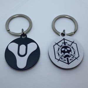 Op de afbeelding: Twee sleutelhangers met metalen ringen. De eerste sleutelhanger heeft een wit en zwart logo met een symbool dat lijkt op een gestileerde letter "V". De tweede sleutelhanger heeft een witte achtergrond met een zwart schedel- en kruisbeenderontwerp in een zeshoek.