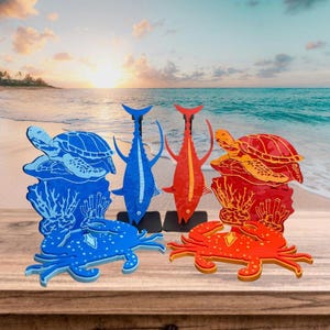 May include: Wooden sea creature sculptures in blue, red, and orange. The collection includes a turtle, fish, and crab, each with intricate details. The sculptures are displayed on a wooden surface with a beach and ocean backdrop.