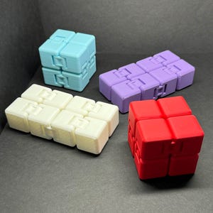 May include: Four fidget cubes in different colors: light blue, white, purple, and red. The cubes are made of plastic and have a variety of textures.