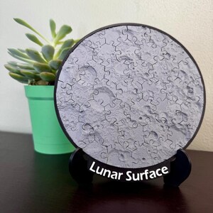Puede incluir: Un rompecabezas circular con un diseño de superficie lunar gris y texturizada con piezas entrelazadas visibles. El rompecabezas está montado sobre un soporte negro con el texto "Lunar Surface" impreso en blanco en la base frontal.