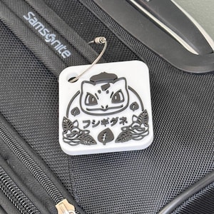 May include: White luggage tag with a black and white illustration of a Pokemon character. The character is surrounded by leaves and the text "フシギダネ" is written in Japanese.