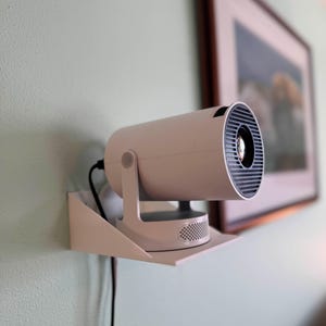 May include: A white projector mounted on a white shelf against a light green wall. The projector has a cylindrical body with a black front and is connected to a black power cord. A framed picture hangs in the background.