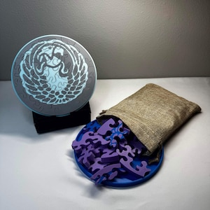 May include: A circular wooden jigsaw puzzle with a grey and light blue design, alongside a blue tray filled with purple pieces. A burlap bag is open, spilling pieces onto the tray. The puzzle is in a round, light blue box.