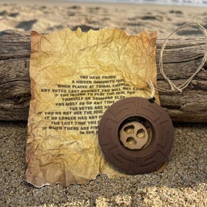 May include: A brown pendant with a beige center, hanging on a natural fiber cord, rests on a sandy surface next to a piece of aged paper with text. The paper reads "YOU HAVE FOUND A HIDDEN IMMUNITY IDOL..."