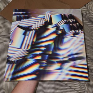 May include: A square print with an abstract design featuring a blend of geometric shapes and vibrant colors. The artwork showcases a mix of blues, greens, and reds, creating a dynamic and visually engaging composition. The print is held in a cardboard box.