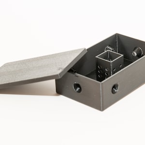 May include: A gray plastic box with a lid, featuring a small, square, open compartment inside. The box has several small holes on the sides and top.