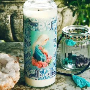 May include: A white candle with a blue and white patterned design featuring a religious image of a woman in a blue robe. The candle is in a glass jar and is sitting on a table with a crystal and a glass jar with teal and purple stones.