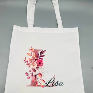 Könnte beinhalten: Weiße Einkaufstasche mit einem Blumendesign mit einem rosafarbenen und goldenen Buchstaben L und dem Namen Lisa in schwarzer Schrift.