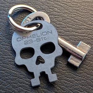 May include: A silver skull keychain with a key attached. The skull has the words "OPHELION 23-61t" engraved on it.