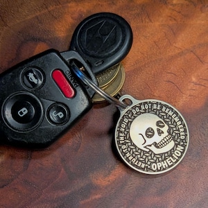 May include: A silver-colored keychain pendant with a skull design and the words "RETURN TO THE VOID, DO NOT BE REMEMBERED, OPHELION" is attached to a car key fob. The fob is black with several buttons and a red "PANIC" button.