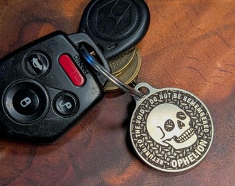 May include: A silver-colored keychain pendant with a skull design and the words "RETURN TO THE VOID, DO NOT BE REMEMBERED, OPHELION" is attached to a car key fob. The fob is black with several buttons and a red "PANIC" button.