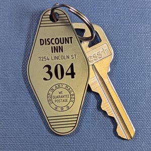May include: A vintage-style key tag from the Discount Inn, featuring the address 7254 Lincoln St and room number 304. The tag is attached to a key with the word "ess+" on it. The tag also has the text "Drop in any mailbox, we guarantee postage."