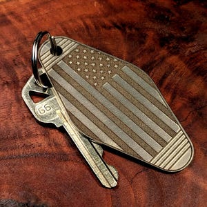 May include: A metal key tag with an engraved American flag design. The tag is attached to a silver key ring and rests on a wooden surface. A silver key with the number 66 is also present.