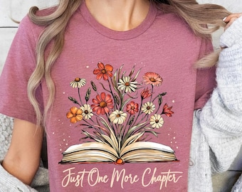 May include: A mauve t-shirt featuring a floral design emerging from an open book. The flowers are in shades of orange, red, and white. The text "Just One More Chapter" is written in a script font below the book.