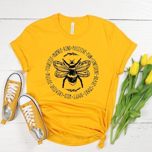 May include: Mustard yellow t-shirt with a black bee graphic and inspirational words. The shirt is paired with matching sneakers and a bouquet of yellow tulips, all set against a white wood background.