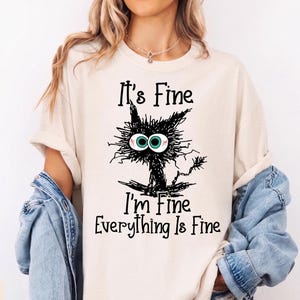 May include: An ivory t-shirt featuring a black scribbled illustration of a wide-eyed cat with teal irises. The text printed on the shirt reads: It's Fine, I'm Fine, Everything Is Fine. The shirt has short sleeves and a crew neckline.