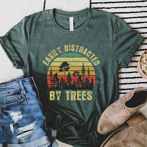 May include: Green t-shirt with the text "EASILY DISTRACTED BY TREES" in a circular design with a sunset and tree silhouette. The shirt is paired with denim shorts, brown shoes, and a small potted cactus.