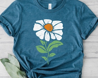 May include: Teal-colored t-shirt featuring a large white daisy with an orange center and green stem and leaves. The shirt is made of soft material and is suitable for casual wear. The design is a floral graphic.