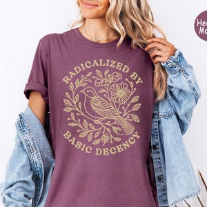 May include: A heather maroon t-shirt featuring a gold-colored graphic of a bird surrounded by floral branches. The text RADICALIZED BY BASIC DECENCY is printed in a circular arrangement around the central bird and floral illustration.