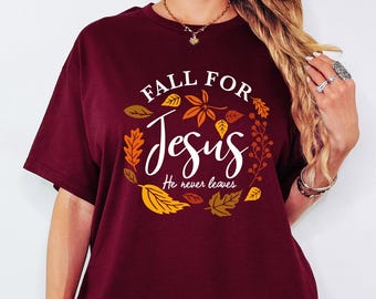 May include: A maroon short-sleeved t-shirt featuring a graphic of autumn leaves in shades of orange, yellow, and brown. The text reads "FALL FOR Jesus He never leaves" in white and script lettering. The shirt is paired with grey ribbed shorts.