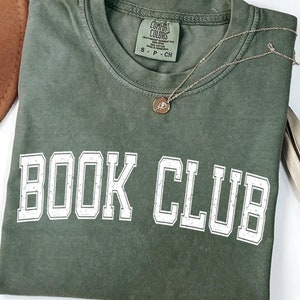 May include: A green t-shirt with the words "BOOK CLUB" printed in white on the front. The shirt is made of hemp and has a relaxed fit.