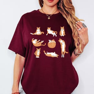 May include: Maroon t-shirt featuring a playful design of orange and white cats in various poses. The cats are arranged in a grid pattern. The shirt has short sleeves and a round neckline. The word "Maroon" is printed on the bottom right.