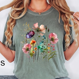 May include: A moss green short-sleeved t-shirt featuring a colorful graphic print of various wildflowers in shades of pink, purple, blue, yellow, and green. The shirt has a crew neckline and a relaxed fit, with the word Moss printed in black.