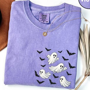 May include: A violet short-sleeved t-shirt with a crew neckline featuring a graphic print of three white ghosts and multiple black bats. The shirt has a Comfort Colors brand label on the inside collar and a circular violet tag labeled VIOLET.