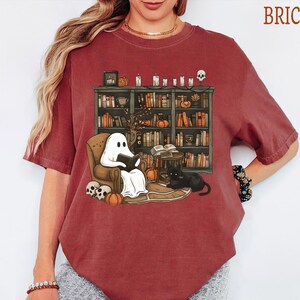 May include: A brick-colored short-sleeve t-shirt featuring a graphic print of a ghost reading a book in an armchair. The scene includes a bookshelf filled with books, pumpkins, skulls, candles, and a black cat. The word BRICK is printed in the corner.