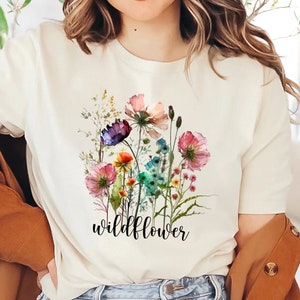 May include: Cream-colored t-shirt featuring a watercolor floral design with pink, purple, blue, and orange flowers. The word "wildflower" is printed in black script below the flowers. The shirt has short sleeves.