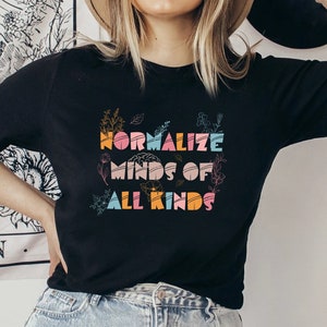 May include: Black t-shirt with the text "NORMALIZE MINDS OF ALL KINDS" in colorful, stylized letters. The design includes floral and botanical elements. The shirt is a classic crew neck style.