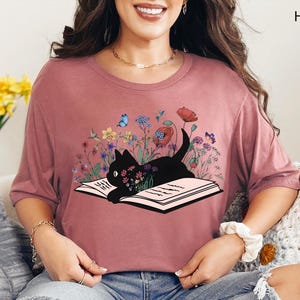 May include: Heather mauve t-shirt featuring a black cat lying on an open book, surrounded by colorful flowers and butterflies. The shirt has short sleeves and a round neckline. The text "HEATHER MAUVE" is printed on the upper right.