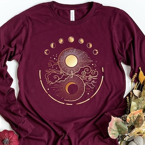 May include: Maroon long-sleeve shirt with a gold celestial design. The design features a sun, moon, clouds, and phases of the moon. The shirt is made of a soft material and is a comfortable fit.