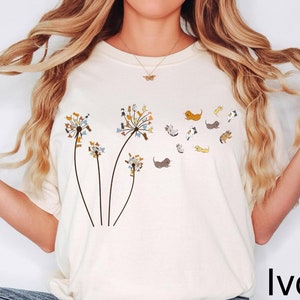 May include: A woman wearing a cream colored t-shirt with a dandelion graphic featuring small dogs blowing in the wind. The text "Ivory" is in the bottom right corner of the image.