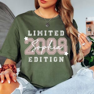 May include: Military green t-shirt with the text "LIMITED 2008 Sophia EDITION" in a pink and white design. The shirt is a crew neck and is worn with blue jeans.