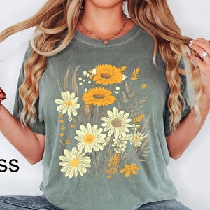 May include: Moss green t-shirt featuring a floral design with yellow sunflowers and white daisies. The graphic includes various wildflowers and the word "Moss" in the lower left corner. The shirt has short sleeves and a relaxed fit.