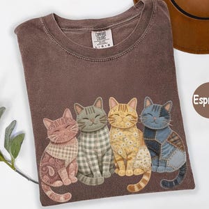 May include: Espresso-colored t-shirt featuring four appliqued cats with unique patterns and colors. The cats are sitting side-by-side. The shirt has a crew neck and a tag that says "Comfort Colors".
