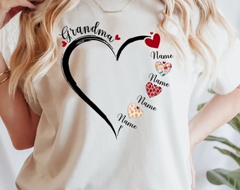 May include: Cream-colored t-shirt featuring a large black heart design with the word "Grandma" above it. The heart has several smaller hearts with the word "Name" printed on them. The shirt is a casual, everyday wear item.