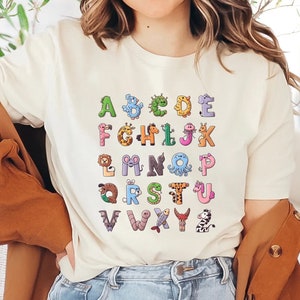 May include: White t-shirt with a colorful alphabet design featuring animals. Each letter is formed by a different animal, such as a giraffe, a penguin, a worm, and a zebra.